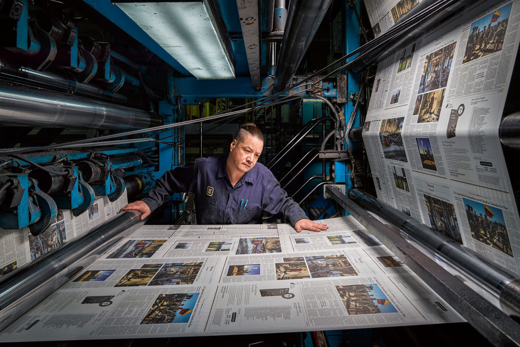 Homme en tenue de travail surveillant la sortie papier d’une rotative dans une imprimerie, inspectant des pages de journaux fraîchement imprimées dans un environnement industriel.