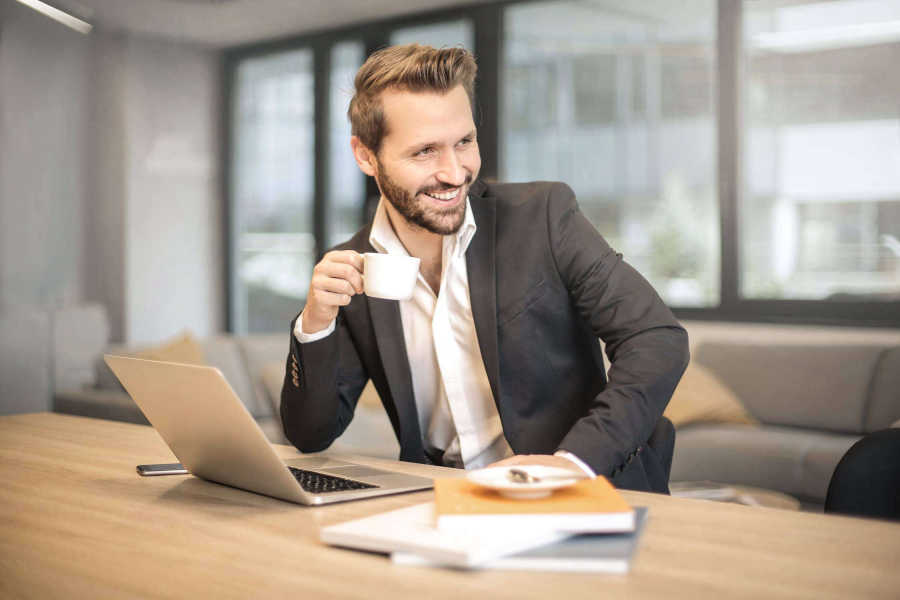 Homme souriant en costume, tenant une tasse de café devant son ordinateur portable dans un bureau lumineux — image symbolisant le dynamisme professionnel et l’épanouissement au travail.